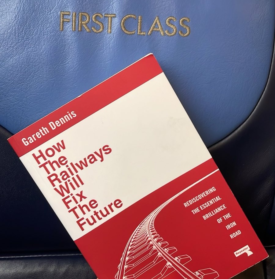 A photograph of Gareth Dennis' 'How The Railways Will Fix The Future: Rediscovering the essential brilliance of the Iron Road'. It's a red covered book with a white title band, and a curved darawing of a rail stretching off into the distance.  It is held up at a 45 degree angle below an embroidered seat cover reading 'First Class'. The seat cover is a light blue with silver embroidery. The cover is surrounded by a much darker blue on the outer edges.